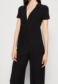 Svart jumpsuit med korta ärmar, djup V-ringning, vriden frontdetalj och vida byxor. Tillverkad av ett mjukt, stretchigt material för komfort.