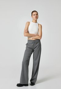 Witte geribbelde tanktop gecombineerd met grijs gestreepte wijde broek en zwarte plateauschoenen. Model staat met de armen gekruist tegen een effen achtergrond.