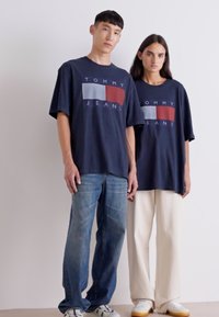 Camisetas de gran tamaño en azul marino con el gráfico "TOMMY JEANS" en rojo y azul claro. Usadas con jeans claros y pantalones de pierna ancha en crema.