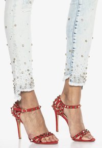 Czerwone zamszowe sandały na wysokim obcasie z złotymi ćwiekami. Połączone z jasnoniebieskimi dżinsami z ozdobnymi mankietami i postrzępionymi brzegami.