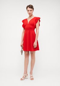 Robe rouge en ligne A avec des manches courtes à volants et un décolleté en V. Tissu lisse avec une jupe évasée. Modèle tenant une pochette argentée et portant des sandales blanches.