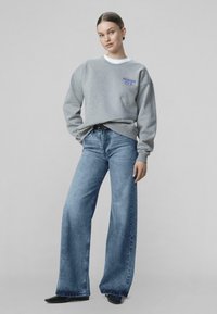 Jauna moteris, apsirengusi pilku džemperiu su mėlynu logotipu, aukšto liemens plačiomis kojomis mėlynais džinsais ir juodais smailiais batais, stovinti prie neutralaus fono.