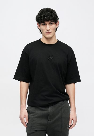 Black oversized t-shirt with short sleeves, featuring a subtle embroidered design on the front. Soft fabric with a relaxed fit, paired with dark pants.