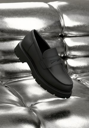 Musta nahkakengät, joissa on kohotettu etuosa, teksturoitu pinta ja kova musta pohja, kiiltävän metallitaustan edessä.