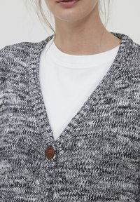 Cardigan en tricot gris et blanc avec un col en V, équipé d'une fermeture à boutons en bois et d'un motif texturé. Porté par-dessus une chemise blanche.