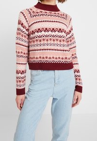 Maglione lavorato a maglia di un rosa chiaro con accenti borgogna, caratterizzato da un motivo geometrico e colletto e polsini a coste. Abbinato a jeans azzurri chiari.