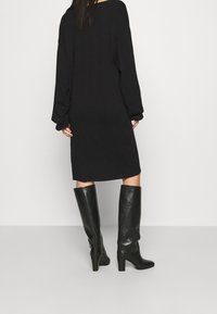 Kvinna klädd i en lös svart långärmad klänning och knähöga svarta läderstövlar med klack, stående mot en ljus enfärgad bakgrund.