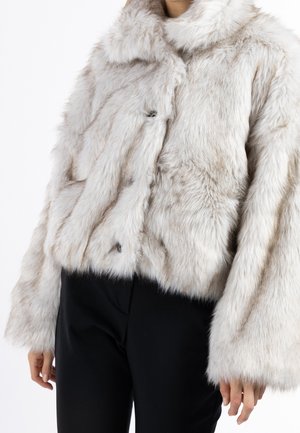 Frau trägt eine hellbeige Jacke aus Kunstpelz mit Kragen und langen Ärmeln, kombiniert mit schwarzer Hose vor weißem Hintergrund.