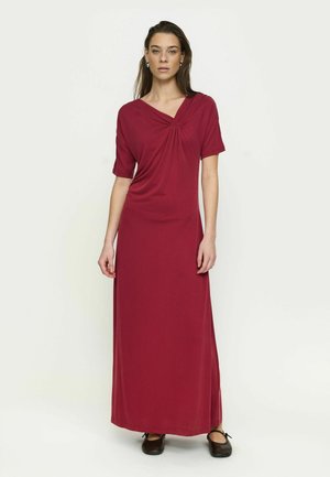 Kvinde står iført en gulvlængde mørkerød kjole med korte ærmer og en rynket detalje ved udskæringen, kombineret med sorte flade sko.