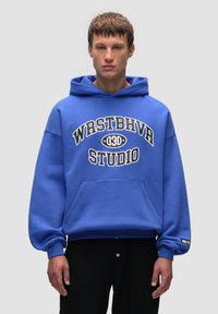 Niebieska bluza z kapturkiem, wykonana z miękkiego materiału, z kieszenią z przodu, z wyraźnym białym napisem "WRSTBHVR STUDIO" oraz małym logo na rękawie.