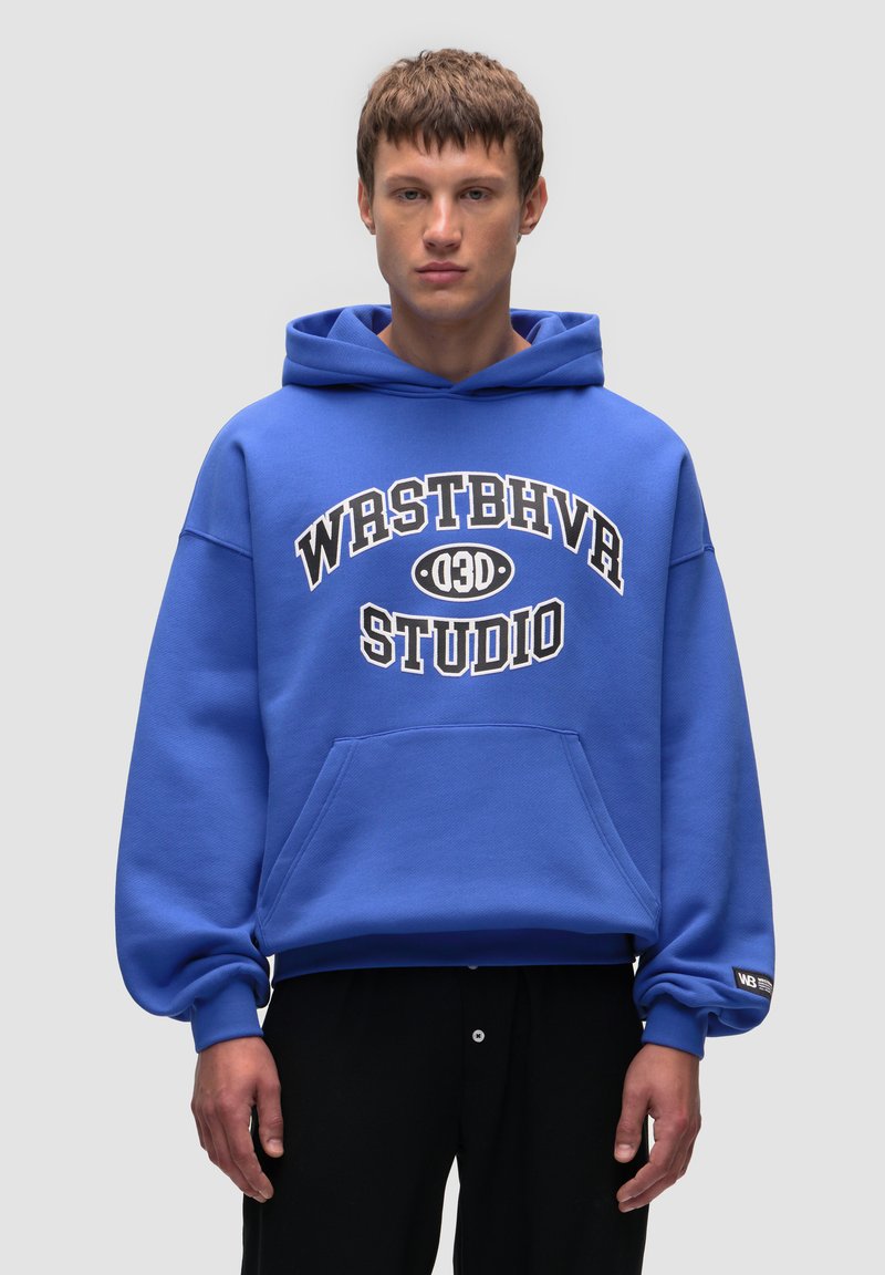 Niebieska bluza z kapturkiem, wykonana z miękkiego materiału, z kieszenią z przodu, z wyraźnym białym napisem "WRSTBHVR STUDIO" oraz małym logo na rękawie.