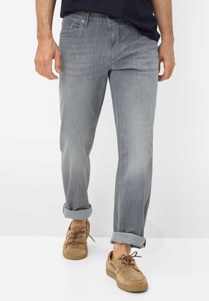 Mand iført lysegrå opsmøgede jeans og brune ruskindssnøresko, gående på en ensfarvet hvid baggrund.