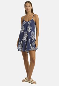 Modelo vistiendo un vestido floral azul marino con tirantes delgados, de pie con confianza con las manos a los lados, combinado con sandalias beige.