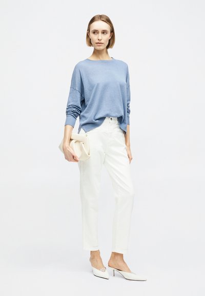 Γυναίκα που φοράει ανοιχτό μπλε πουλόβερ και άσπρο παντελόνι, κρατώντας μια μπεζ τσάντα χειρός, στέκεται μπροστά σε απλό λευκό φόντο.