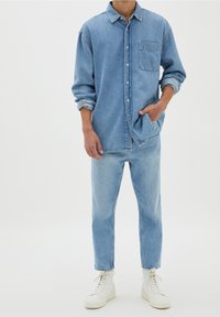 Camisa de mezclilla azul claro con botones, bolsillos y mangas largas, combinada con jeans azul claro a juego y zapatillas altas blancas.