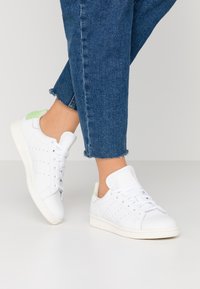 Person som bär vita sneakers med gröna hältappar och blå beskurna jeans, stående mot en enhetlig ljus bakgrund.