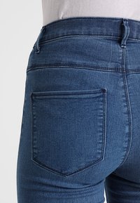 Jeans in denim blu a vita media con una vestibilità slim, dotati di tasche posteriori, cuciture sottili e una texture liscia.