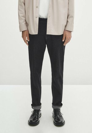 Homme portant un pantalon noir retroussé, des chaussures noires à lacets, une chemise blanche et une veste beige à boutons, debout sur un sol blanc.
