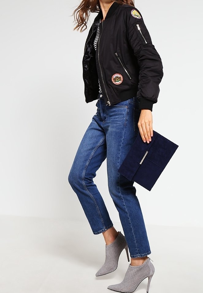 Nainen, jota on yllään musta bomber-takki, siniset farkut ja harmaat korkeat nilkkurit, pitää kädessään tummansinistä käsilaukkua.