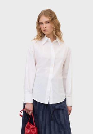 Nuori nainen, jolla on aaltoilevat vaaleat hiukset, pukeutunut valkoiseen napitettavaan paitaan ja tummaan raidalliseen hameeseen, pitää pientä punaista käsilaukkua sivullaan.