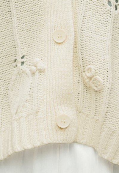 Κρεμ πλεκτή ζακέτα με υφασμάτινα λουλουδένια μοτίβα και δύο εμφανή κουμπιά σε ραβδωτό τελείωμα.