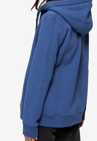 Person wearing a blue hooded sweatshirt with long sleeves and black pants, shown from the side against a plain background.