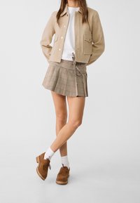 Beige wollen jas met knopen aan de voorkant, wit T-shirt, geruite minirok en bruine suède schoenen. De rok heeft een strikceintuur en plooien.