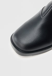 Bota de couro preto com acabamento texturizado, apresentando bico arredondado e costura branca contrastante ao longo da costura superior. Sola preta plana.