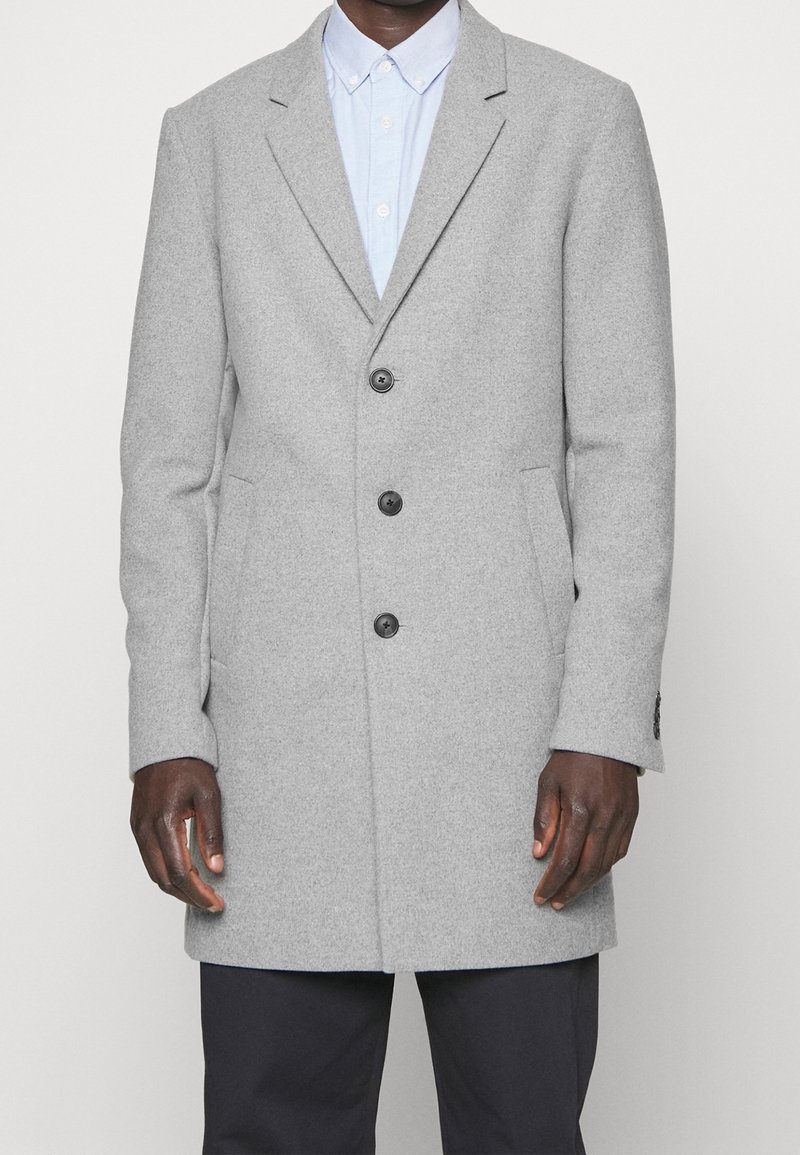 Mężczyzna ubrany w jasnoszary wełniany płaszcz z trzema czarnymi guzikami, na niebieskiej koszuli z kołnierzykiem i czarnych spodniach, stoi na jednolitym tle.