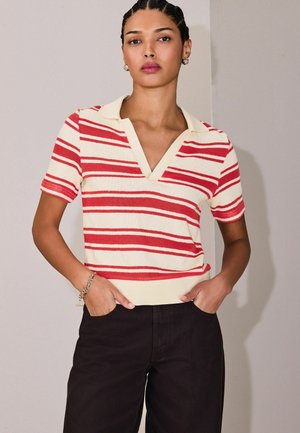 Frau mit geflochtenem Haar, trägt ein creme- und rotgestreiftes kurzärmeliges Strickoberteil und schwarze Hosen, Hände in den Taschen, vor einer neutralen Wand.