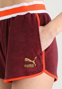 Bordové froté šortky Puma s oranžovým lemem a logem, elastický pás s bílými a oranžovými pruhy, ruka v kapse.