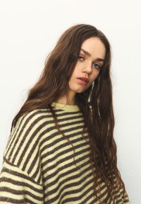 Nadmerný pletený sveter s prestriedanými hnedými a krémovými pruhmi, vyrobený z mäkkého materiálu, s širokým okrúhlym výstrihom a voľným strihom.