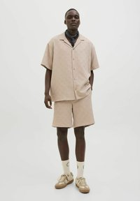 Cămașă cu mânecă scurtă, model bej, asortată cu pantaloni scurți din același material, realizată din țesătură ușoară. Accesoriile includ un fular negru și adidași bej.