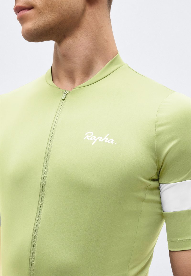Nahaufnahme einer Person, die ein hellgrünes Radtrikot mit Reißverschluss und dem "Rapha"-Logo auf der Brust sowie einem weißen Streifen am Ärmel trägt.