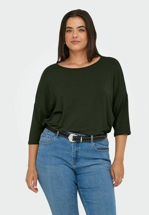 Mujer con cabello largo y oscuro, vistiendo una blusa suelta de color verde oscuro, jeans azul claro y un cinturón negro con tachuelas, de pie frente a un fondo liso.