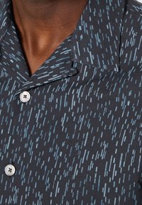 Zbliżenie na ciemną koszulę w wzory z jasnoniebieskimi abstrakcyjnymi liniami i białymi guzikami na szyi i górnej części klatki piersiowej osoby.