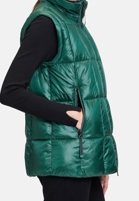 Zelená prošívaná vesta bez rukávů s vysokým límcem, s boční kapsou na zip, má lesklý povrch a prošívanou texturu.