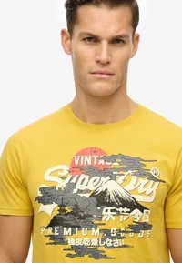 Camiseta de algodón amarilla con un estampado gráfico de una escena montañosa con nubes y texto. Cuello redondo y mangas cortas.