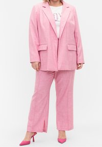 Femme portant un tailleur rose avec un pantalon à jambes larges, un blazer, un tee-shirt blanc à motif floral et des escarpins roses à bout pointu, debout devant un fond blanc.