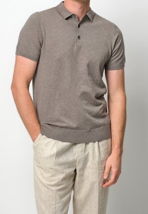 Homme portant un polo taupe à manches courtes et un pantalon beige clair, une main dans la poche et l’autre détendue le long du corps.