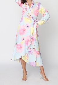 Femme portant une robe portefeuille fleurie pastel avec un ourlet à volants et des manches longues, posant avec une main sur la hanche et un bras levé.