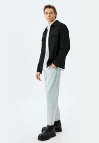 Chaqueta negra con dos bolsillos delanteros, llevada sobre una camisa blanca; jeans de mezclilla azul claro y botas chunky negras; fondo neutro.