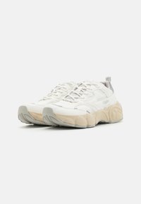 Weiße Sportschuhe aus Mesh und synthetischen Materialien. Sie verfügen über eine beigefarbene Gummisohle, subtile graue Akzente und texturierte Designelemente.