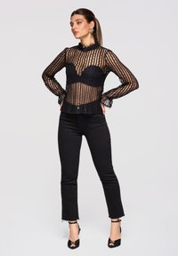 Femme debout, mains sur les hanches, portant un haut en dentelle noire transparente, un pantalon court noir et des talons ouverts noirs, sur un fond uni.
