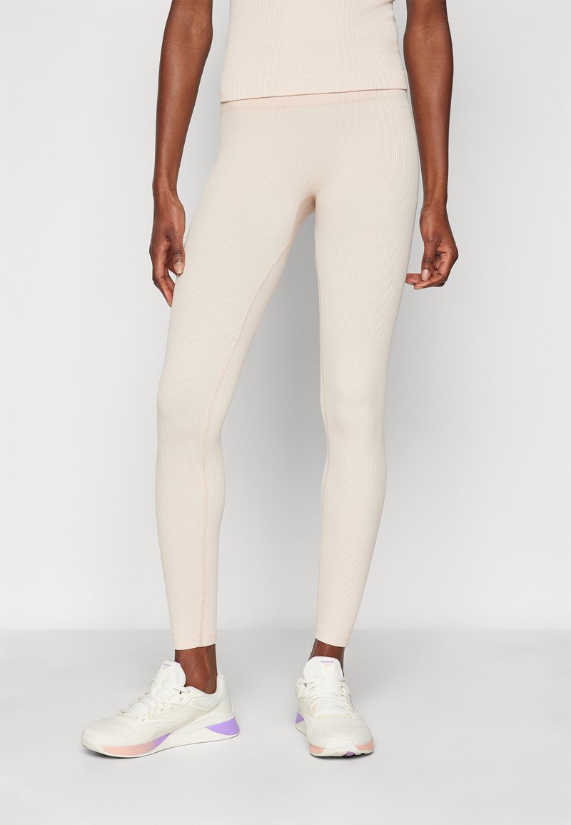 Beige bordázott leggings, testhezálló szabással és kontrasztos varrásokkal, fehér sportcipővel párosítva, lila és őszibarack színekkel kiemelve.
