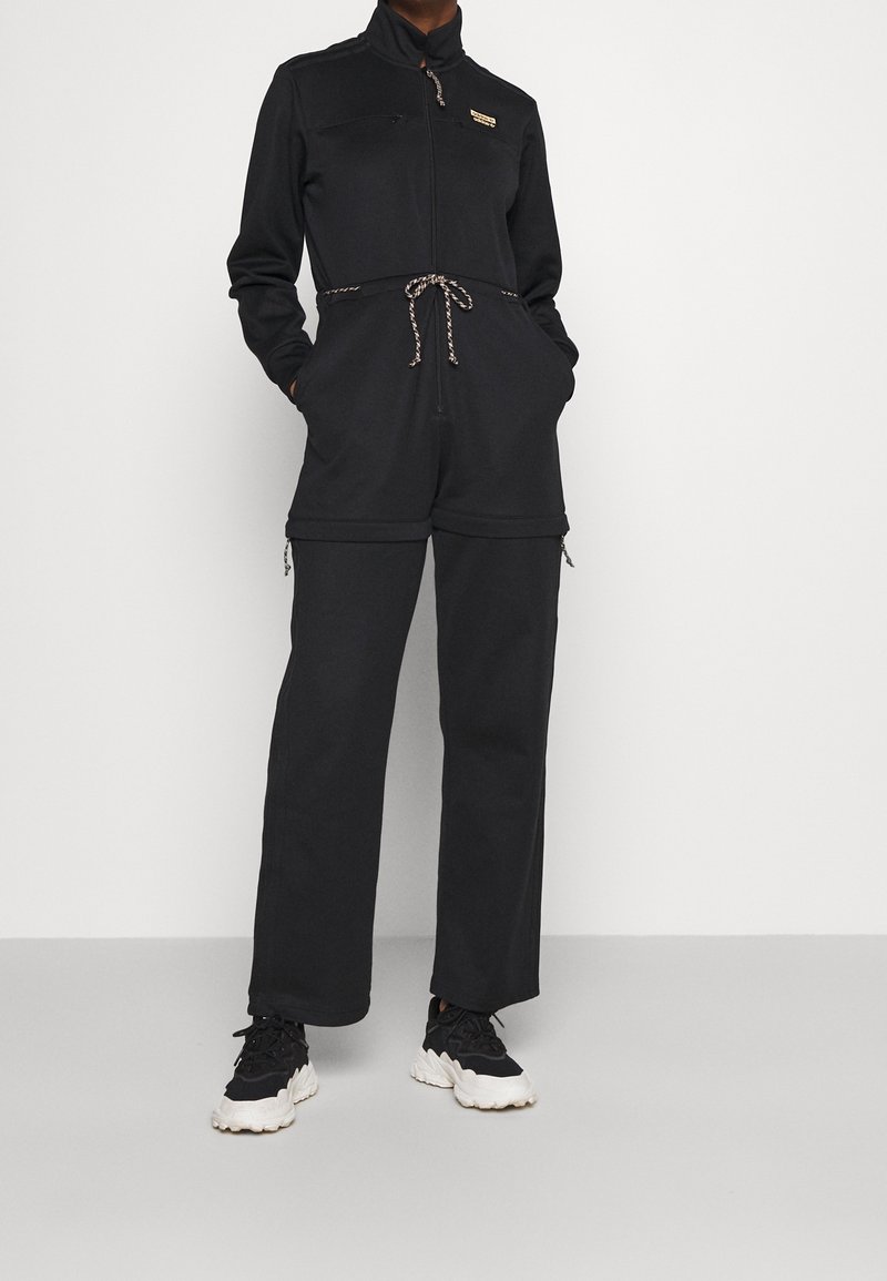 Černý overal vyrobený z měkké tkaniny se zipem vpředu, stahovací pasem a dvěma předními kapsami. Obsahuje žebrované manžety a malé logo.