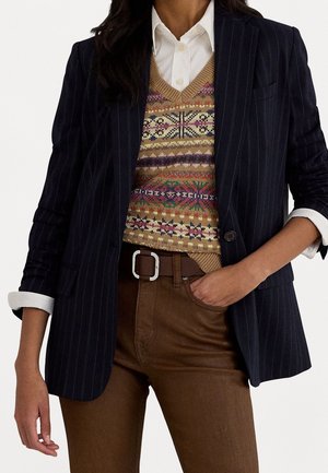 Persona que lleva una camisa blanca, un chaleco tejido con un patrón multicolor, un blazer azul marino con rayas diplomáticas y pantalones marrones con cinturón, con la mano en el bolsillo.