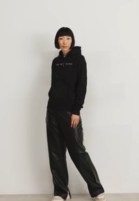 Sudadera negra con texto blanco "TOMMY JEANS", combinada con pantalones de cuero sintético negro y zapatillas crema, todo sobre un fondo neutro.