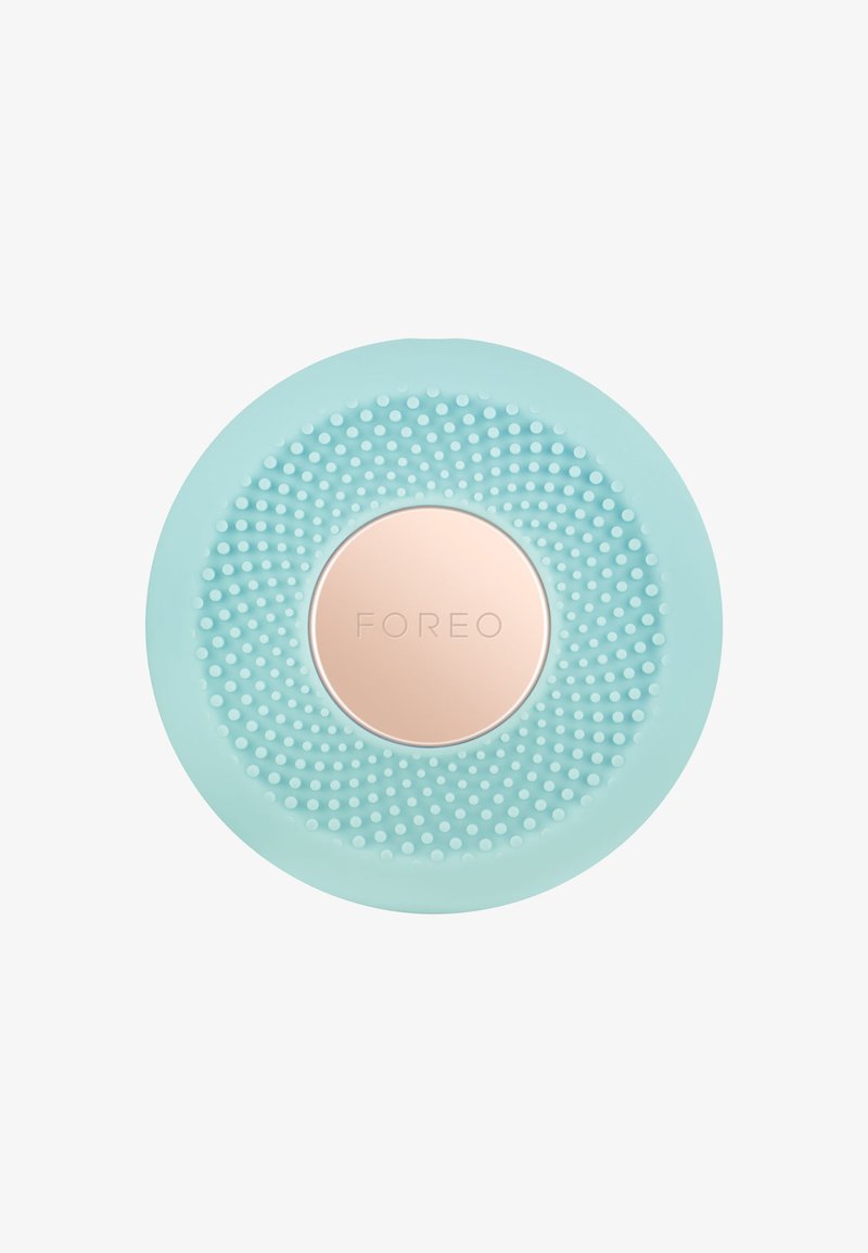 Sirkulær renseenhet i mintgrønn silikon, med en teksturert overflate av små prikker og en sentral logo plate i roségull.