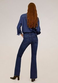 Jeans acampanados de mezclilla oscura con cintura alta, diseño de cinco bolsillos y dobladillo deshilachado. Combinados con una camisa de mezclilla de manga larga a juego.